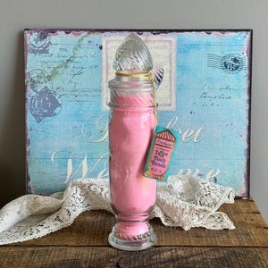 May include: A clear glass apothecary jar filled with bright pink bath beads, topped with a decorative twisted glass lid. A small rectangular tag attached with a yellow string reads Proclaim Foaming Bath Beads Net Wt 2 oz in pink and blue.