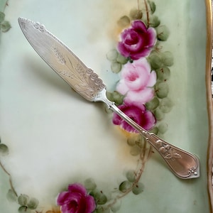 May include: A silver-toned metal butter knife featuring an ornate handle with embossed floral details and a blade decorated with an intricate engraved wheat pattern. The knife is positioned diagonally across a pale green surface with painted roses.