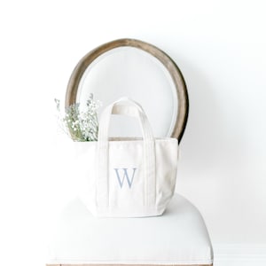 May include: A small, off-white canvas tote bag with a light blue letter "W" is displayed on a white chair. The bag has two handles and is filled with dried flowers. The chair has a wooden frame and is set against a white wall.