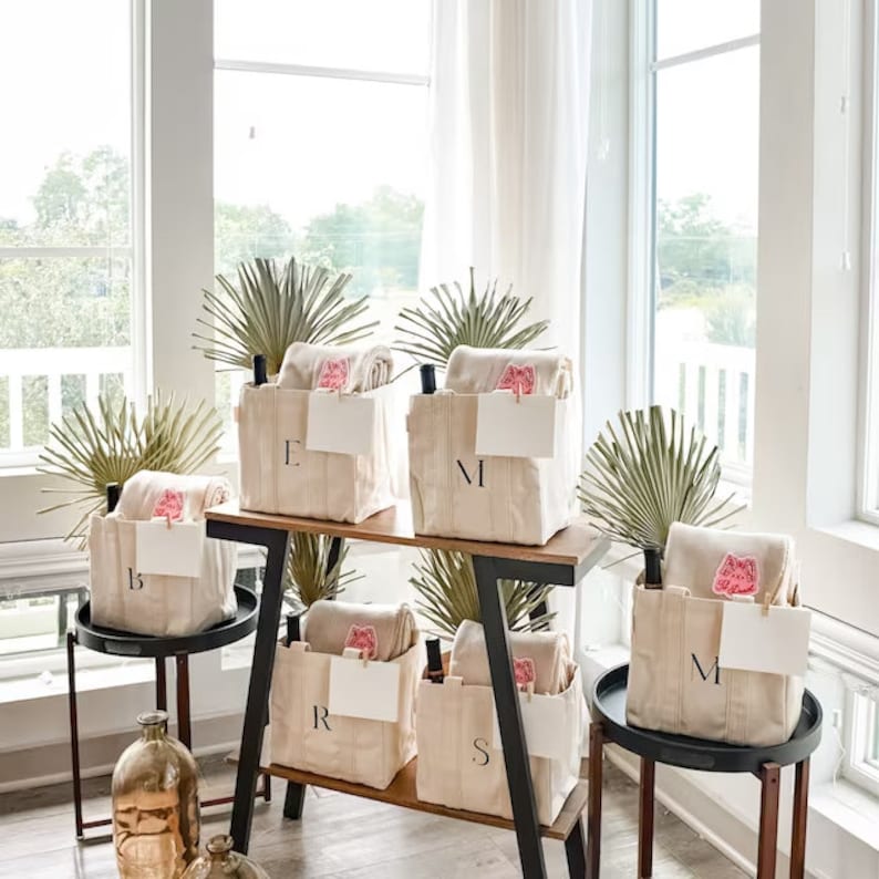 Five natural canvas tote bags with pink and white blankets inside. Each bag has a different letter monogrammed on the front. The bags are arranged on a wooden shelf with a black metal frame.