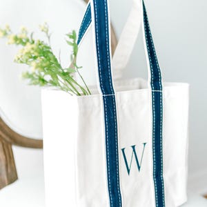 A natural canvas tote bag with navy blue straps and a navy blue hand painted letter "W" on the front.