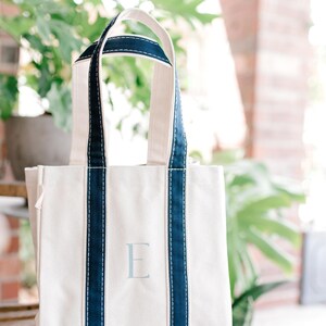 A natural canvas tote bag with blue striped handles and a light blue hand painted letter "E" on the front. The bag is sitting on a wooden rocking chair.