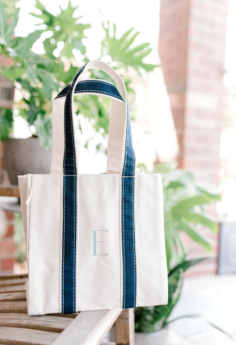 A natural canvas tote bag with blue striped handles and a light blue hand painted letter "E" on the front. The bag is sitting on a wooden rocking chair.