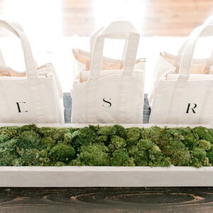 Three natural canvas tote bags with black hand painted letters 'L', 'S', and 'R' on a wooden table with a rectangular white planter filled with green moss.