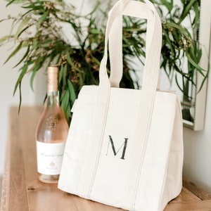 A natural canvas tote bag with a black letter "M" hand painted on the front. The bag has a zipper closure and two handles. A bottle of rose wine is sitting on a wooden table next to the bag.