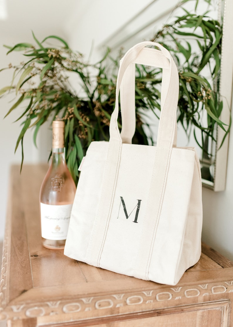 A natural canvas tote bag with a black letter "M" hand painted on the front. The bag has a zipper closure and two handles. A bottle of rose wine is sitting on a wooden table next to the bag.