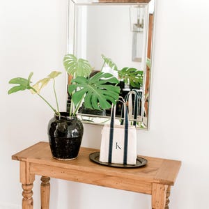 A wooden console table with a black ceramic vase filled with a green monstera plant. A small natural canvas tote bag with blue stripes and the letter "K" hand painted on it sits on a silver tray on the table.