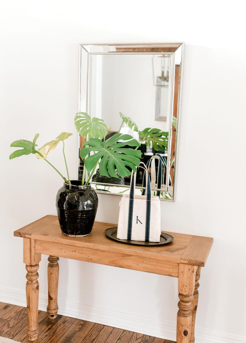 A wooden console table with a black ceramic vase filled with a green monstera plant. A small natural canvas tote bag with blue stripes and the letter "K" hand painted on it sits on a silver tray on the table.