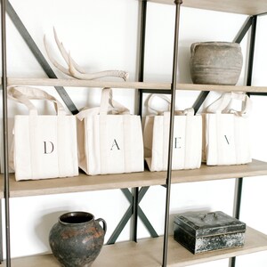 A wooden shelf unit with black metal accents and four shelves. The top shelf has a set of antlers and a brown clay pot. The second shelf has four natural canvas tote bags with the letters D, A, E, and V printed on them. The third shelf has a brown clay pot and a black metal box. The bottom shelf has a book.