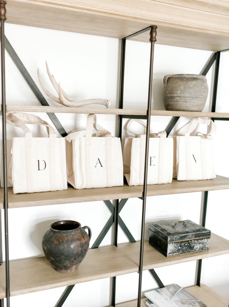 A wooden shelf unit with black metal accents and four shelves. The top shelf has a set of antlers and a brown clay pot. The second shelf has four natural canvas tote bags with the letters D, A, E, and V printed on them. The third shelf has a brown clay pot and a black metal box. The bottom shelf has a book.