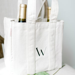 A natural canvas tote bag with a hand painted letter "W" on the front in black. The bag is sitting on a chair.