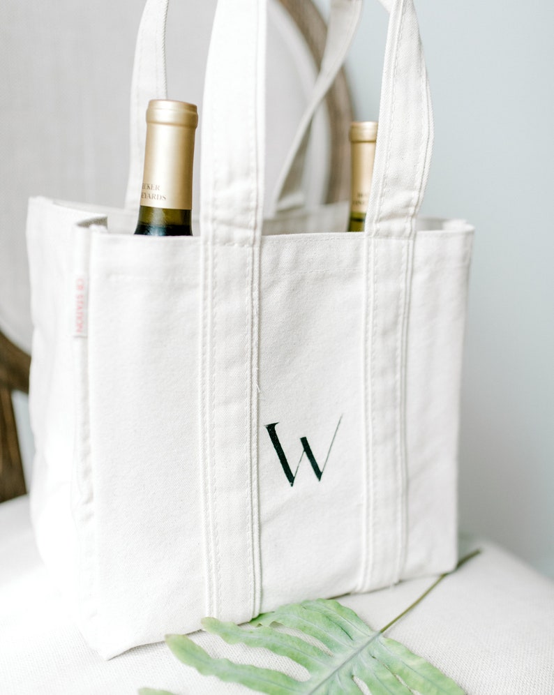 A natural canvas tote bag with a hand painted letter "W" on the front in black. The bag is sitting on a chair.