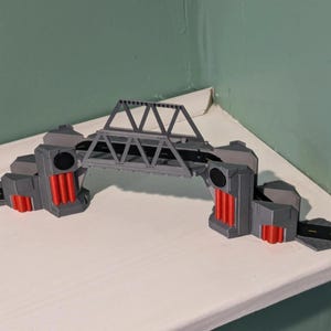 Könnte beinhalten: Eine graue Spielzeugbrücke aus Kunststoff mit einer zentralen Fachwerkstruktur, schwarzen Straßenabschnitten und roten vertikalen zylindrischen Akzenten an den Stützpfeilern. Das Modell enthält integrierte Straßenrampen mit gelben Fahrbahnmarkierungen.
