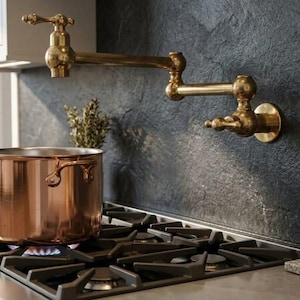 May include: A wall-mounted brass pot filler tap with a double-jointed swing arm and two handles, positioned above a gas hob with a large copper pot. The tap features a metallic gold finish and a classic design with visible joints.