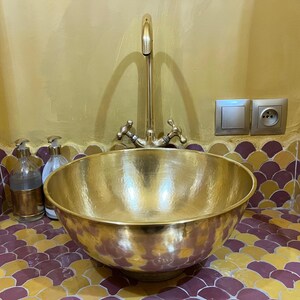 May include: A hammered brass sink basin with a matching faucet. The sink is round and sits on a tiled surface. Soap dispensers are visible to the left. The wall is painted gold.