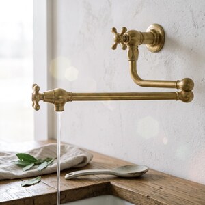 May include: A wall-mounted brass pot filler faucet with a double-jointed swing arm and two cross-style handles. The metallic fixture features a brushed finish and is shown with water flowing from the spout into a sink area below the faucet.