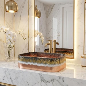 Könnte beinhalten: Ein rechteckiges Waschbecken mit Kupfer-, Gold- und schwarzem Finish. Das Waschbecken steht auf einer Marmorarbeitsplatte, mit goldenen Armaturen und einem Spiegel im Hintergrund. Der Raum ist mit goldenen Akzenten und weißen Orchideen dekoriert.