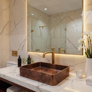 May include: A rectangular copper vessel sink with a hammered texture sits on a white marble countertop with grey veining. A tall, brushed gold faucet is positioned behind the sink. A large rectangular mirror reflects the bathroom interior.