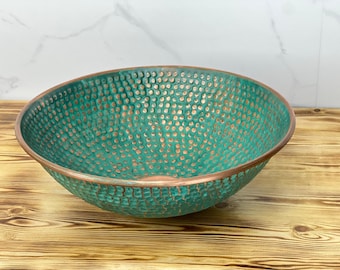 May include: A teal-coloured, hammered copper bowl with a bronze rim. The bowl has a textured surface with small, evenly spaced indentations. The interior has a copper colour. The bowl is set on a wooden surface.