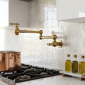 May include: A wall-mounted brass pot filler faucet with a double-jointed swing arm, positioned above a gas stove with a large copper pot. Three glass bottles filled with oil sit on a marble tray next to a small potted green plant.