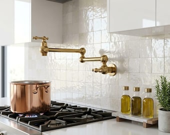 May include: A wall-mounted brass pot filler faucet with a double-jointed swing arm, positioned above a gas stove with a large copper pot. Three glass bottles filled with oil sit on a marble tray next to a small potted green plant.