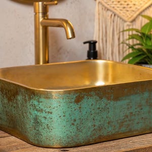 Könnte beinhalten: Ein rechteckiges Waschbecken mit goldener Innenseite und verwitterter türkisfarbener Außenseite. Hinter dem Waschbecken befinden sich ein goldener Wasserhahn und ein schwarzer Seifenspender. Grüne Pflanzen und Handtücher sind im Hintergrund.