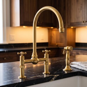 May include: A bridge-style kitchen faucet with a brushed gold finish, featuring a high-arc spout, two cross-handle controls, and a separate side sprayer. The faucet is mounted on a black marble countertop with white veining.