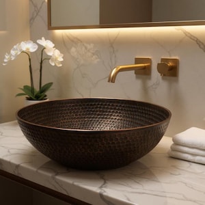 Puede incluir: Un lavabo de color cobre martillado con una superficie texturizada. El lavabo es redondo y se encuentra sobre una encimera de mármol. Un grifo dorado y accesorios a juego están montados en la pared sobre el lavabo. Toallas blancas apiladas a la derecha.