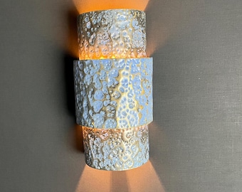 Puede incluir: Aplique de pared cilíndrico de tres niveles con un acabado texturizado en azul claro y dorado. La lámpara emite una luz cálida, creando una iluminación decorativa y funcional. Montada en una pared gris oscuro.