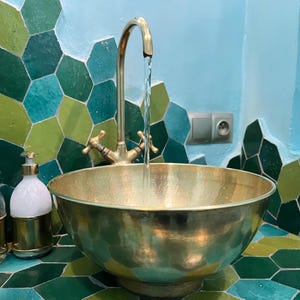 May include: A brass basin sink with a gold faucet, water flowing into the bowl. The sink is set against a wall with green and blue hexagonal tiles. A soap dispenser is visible on the left.