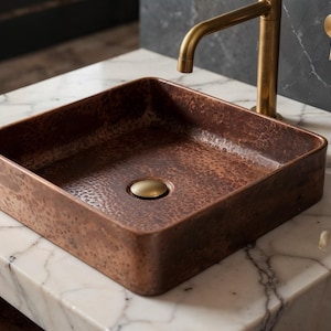 Op de afbeelding: Een vierkante koperen wastafel met een gehamerde textuur staat op een wit marmeren aanrecht. Boven de wastafel zijn een gouden kraan en bijpassende wandarmaturen gemonteerd. De wastafel heeft een gouden afvoer.