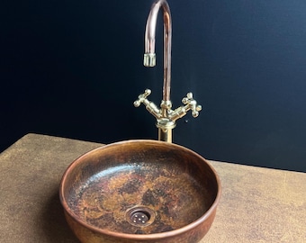 Könnte beinhalten: Ein runder kupferfarbener Waschbecken mit gesprenkelter Oberfläche. Über dem Waschbecken befindet sich ein kupferfarbener Wasserhahn mit goldfarbenen Griffen. Das Waschbecken und der Wasserhahn sind vor einem dunklen Hintergrund platziert.