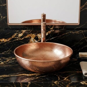 May include: A copper-colored, hammered-metal vessel sink with a matching faucet. The sink is round and sits on a black marble countertop. A mirror and soap dish are visible in the background. A folded linen towel is on the counter.