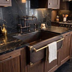 May include: A dark brown hammered copper farmhouse sink with brass corner accents and a horizontal brass towel bar. A light-coloured linen towel hangs from the bar. The sink is set into a dark wood cabinet with black marble countertops.