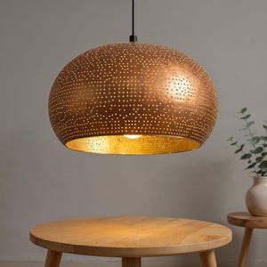 May include: A copper-coloured pendant light with a perforated design hangs above a wooden table. The interior of the light fixture is gold-toned, and a small plant in a white vase sits on a wooden side table. The light fixture is a decorative element.