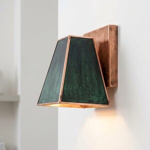 Pode incluir: Luminária de parede com acabamento em cobre. Possui uma sombra trapezoidal verde escura com acabamento em cobre e uma placa traseira de cobre. A luz está acesa, emitindo um brilho quente.