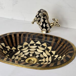 Puede incluir: Un lavabo ovalado con un diseño incrustado decorativo en tonos negros, marrones y crema. Un grifo a juego con un patrón negro y crema y detalles dorados se encuentra encima del lavabo. A la derecha hay un dispensador de jabón con ribete dorado.