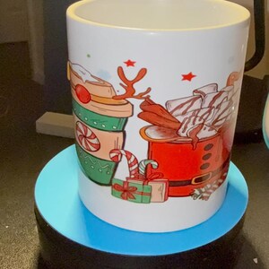 May include: White ceramic mug with a festive Christmas design. The mug features illustrations of a coffee cup, Santa's suit, candy canes, and wrapped gifts. The mug is placed on a blue circular stand.