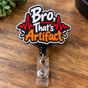 May include: A black badge reel featuring a white and orange graphic design with the text Bro, That's Artifact. The design includes a red heartbeat line across the center. The badge reel has a clear plastic strap with a metal snap button attachment.