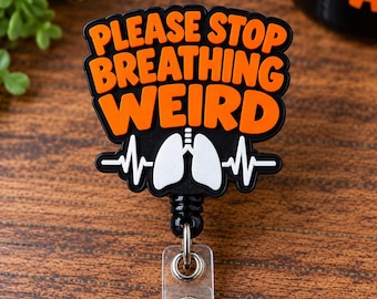 May include: A black badge reel featuring the orange text PLEASE STOP BREATHING WEIRD above a white graphic of human lungs and a heartbeat line. The circular badge holder has a retractable cord and a clear plastic strap with a metal snap button.
