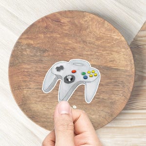 May include: A white and gray Nintendo 64 controller with colored buttons. The controller is on a wooden surface.
