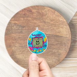 May include: A blue and pink Tamagotchi digital pet toy with a black and white pixelated image of a creature on the screen. The toy has a white border and is surrounded by a colorful, abstract design.