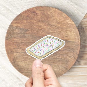 May include: A rectangular sticker resembling a frosted pastry with colorful sprinkles. The sticker has a white frosting base and a tan crust. The sticker is held by a hand, placed on a wooden surface.