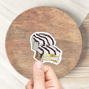 May include: A sticker of two white and brown layered cakes with chocolate frosting and stripes. The cakes are on a wooden plate.
