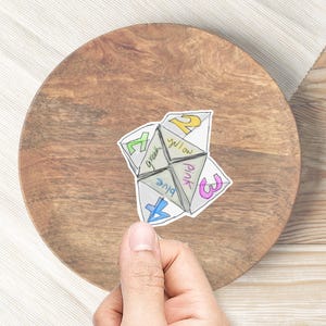 May include: A paper fortune teller with the numbers 1, 2, 3, and 4 on the outside. The inside of the fortune teller has the words "yellow", "pink", "blue", and "green" written on it.