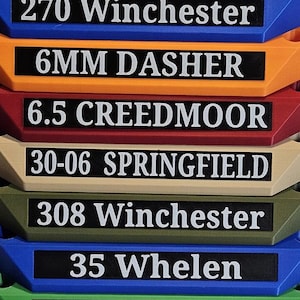 May include: A stack of colorful, angled holders, each labeled with white text on a black background. The labels read: "270 Winchester", "6MM DASHER", "6.5 CREEDMOOR", "30-06 SPRINGFIELD", "308 Winchester", "35 Whelen", and "308 Winchester".