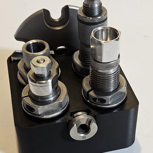 May include: A black metal tool holder with several silver-colored metal components of varying shapes and sizes. The components include cylindrical pieces, nuts, and a curved piece on the left side. The holder is on a white surface.