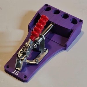 Pode incluir: Um gabarito roxo com uma alça vermelha e uma braçadeira prateada. O gabarito tem quatro furos e o número 16 impresso na lateral. Esta ferramenta é provavelmente usada para projetos de marcenaria ou metalurgia.