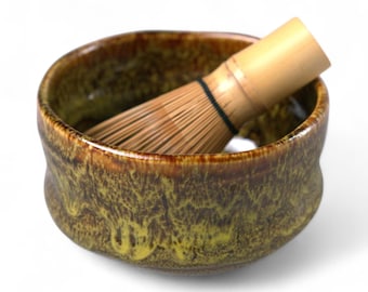 May include: A ceramic matcha bowl with a bamboo whisk. The bowl has a mottled brown and yellow glaze. The whisk is made of light-colored bamboo and is positioned inside the bowl, ready for use.