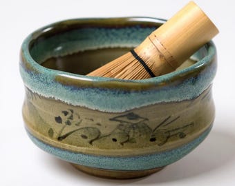 May include: A ceramic matcha bowl with a blue and brown glaze, featuring a bamboo whisk. The bowl has a hand-painted landscape design around the exterior. The whisk is made of bamboo and is used for preparing matcha tea.